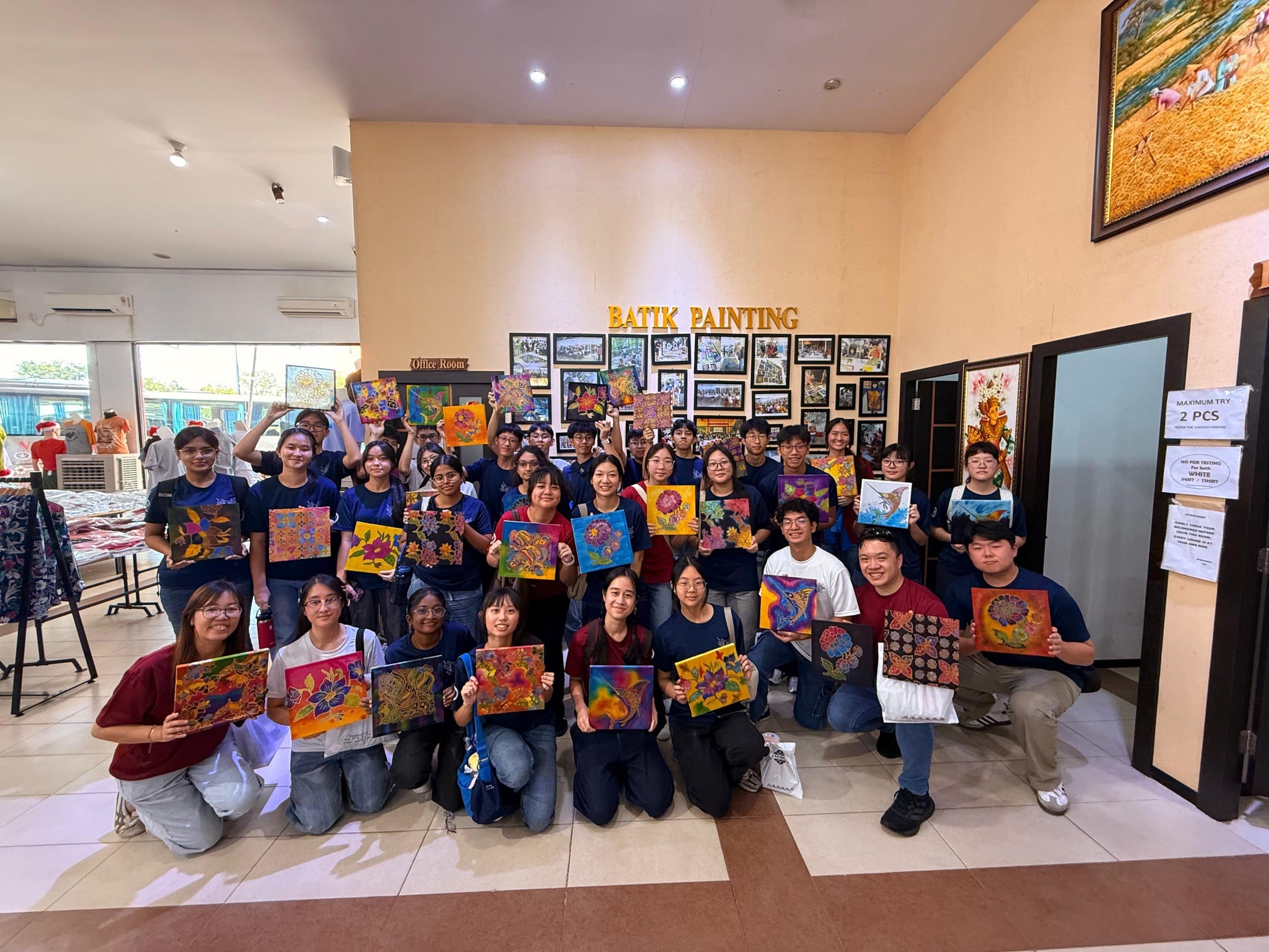 About 35 students and teachers in navy blue t-shirts pose together inside a batik studio in Batam, proudly holding up their completed colourful batik artworks. The words "Batik Painting" are displayed on the wall behind them.